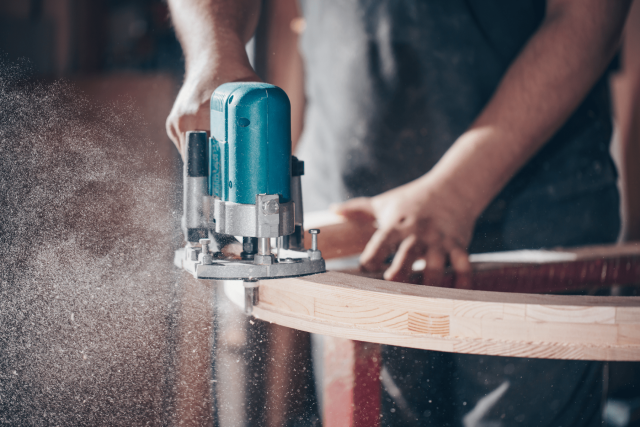 Detailaufnahme: Ein Handwerker schleift den Rahmen eines runden Massivholz-Möbelstücks mit einer Schleifmaschine.