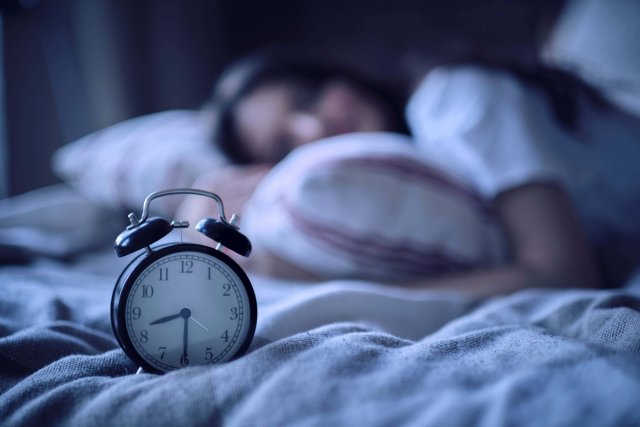 In einem Bett steht ein Wecker der halb neun zeigt. Dahinter sieht man verschwommen eine schlafende junge Frau.