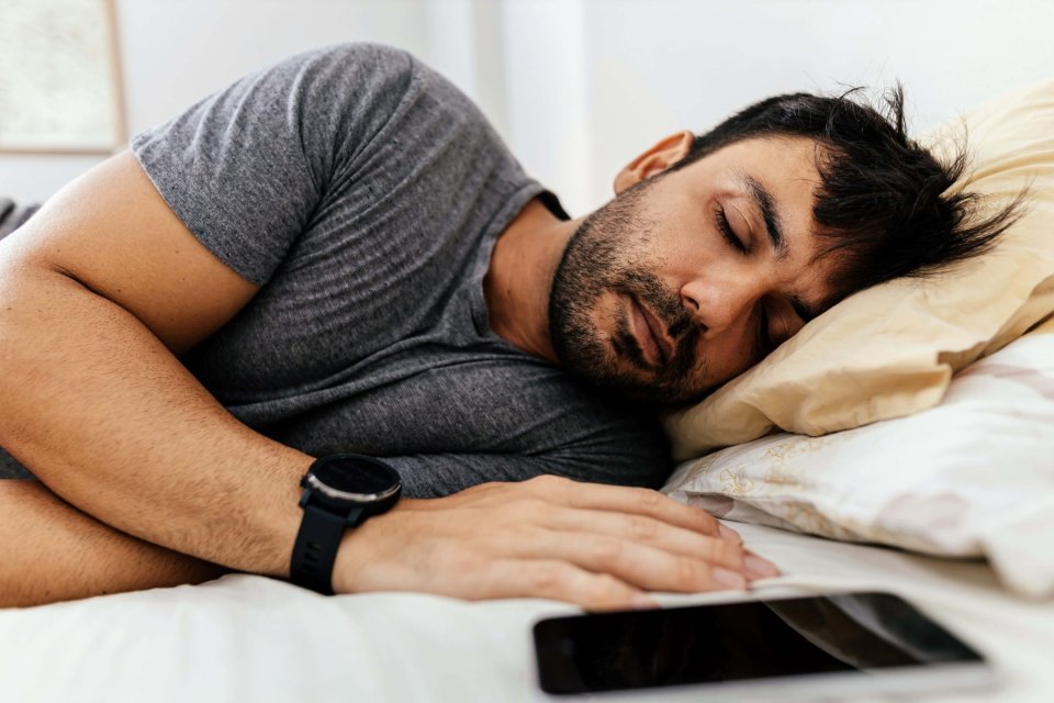 Ein Mann mittleren Alters schläft in einem Bett. Neben ihm liegt ein Handy.