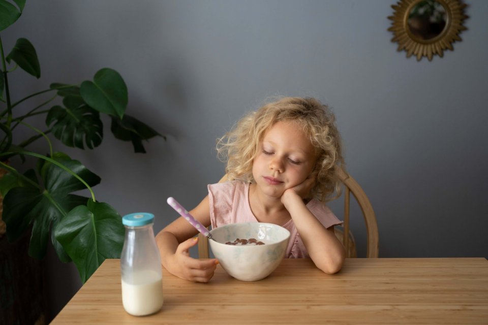 Ein kleines Mädchen mit lockigen blonden Haaren ist über seinem Frühstück eingeschlafen. Es sitzt an einem einfachen Holztisch und hat den Kopf auf den Hand gestützt, die Augen sind geschlossen. Vor ihr steht eine volle Müslischale und eine kleine Flasche Milch.
