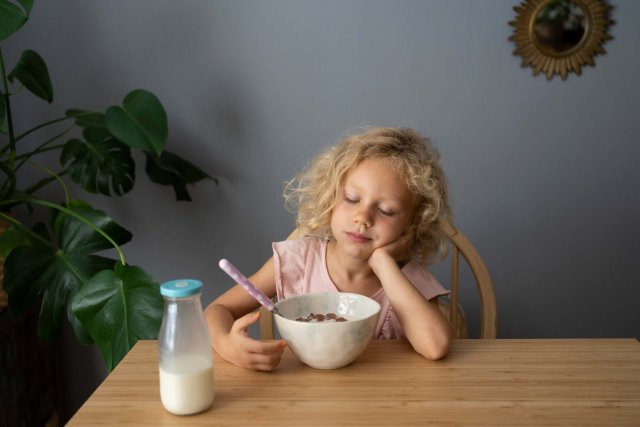 Ein kleines Mädchen mit lockigen blonden Haaren ist über seinem Frühstück eingeschlafen. Es sitzt an einem einfachen Holztisch und hat den Kopf auf den Hand gestützt, die Augen sind geschlossen. Vor ihr steht eine volle Müslischale und eine kleine Flasche Milch.