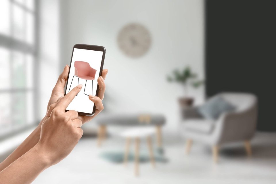 Im Vordergrund sind zwei Hände zu sehen, die ein Smartphone halten. Darauf ist ein pinkfarbener Stuhl zu sehen. Im Hintergrund sieht man verschwommen ein Wohnzimmer.