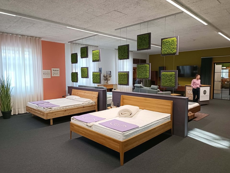Der Ausstellungsraum der Schlafwerkstatt. Im Vordergrund sieht man 2 Massivholzbetten auf denen nur Matratzen und lavendelfarbene Fussschoner liegen. Im Hintergrund ist eine Schlafberaterin und weitere Ausstellungsstücke zu sehen.