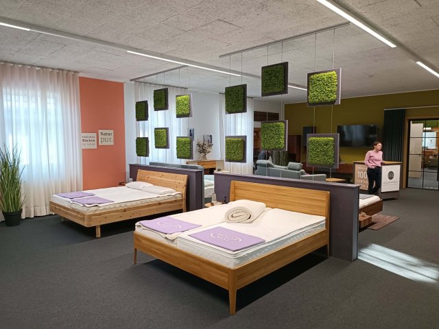 Der Ausstellungsraum der Schlafwerkstatt. Im Vordergrund sieht man 2 Massivholzbetten auf denen nur Matratzen und lavendelfarbene Fussschoner liegen. Im Hintergrund ist eine Schlafberaterin und weitere Ausstellungsstücke zu sehen.
