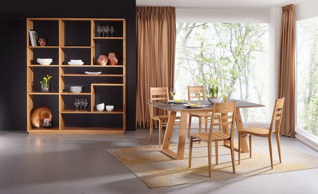 Ein Esszimmer aus MAssivholznöbeln. Links vor einer dunklen Wand steht ein offenes Regal mit unregelmäßigen Fächern. Rechts vor einem bodenhohen Eckfenster ein Esstisch mit blauer Platte und 4 Holzstühlen außen herum.