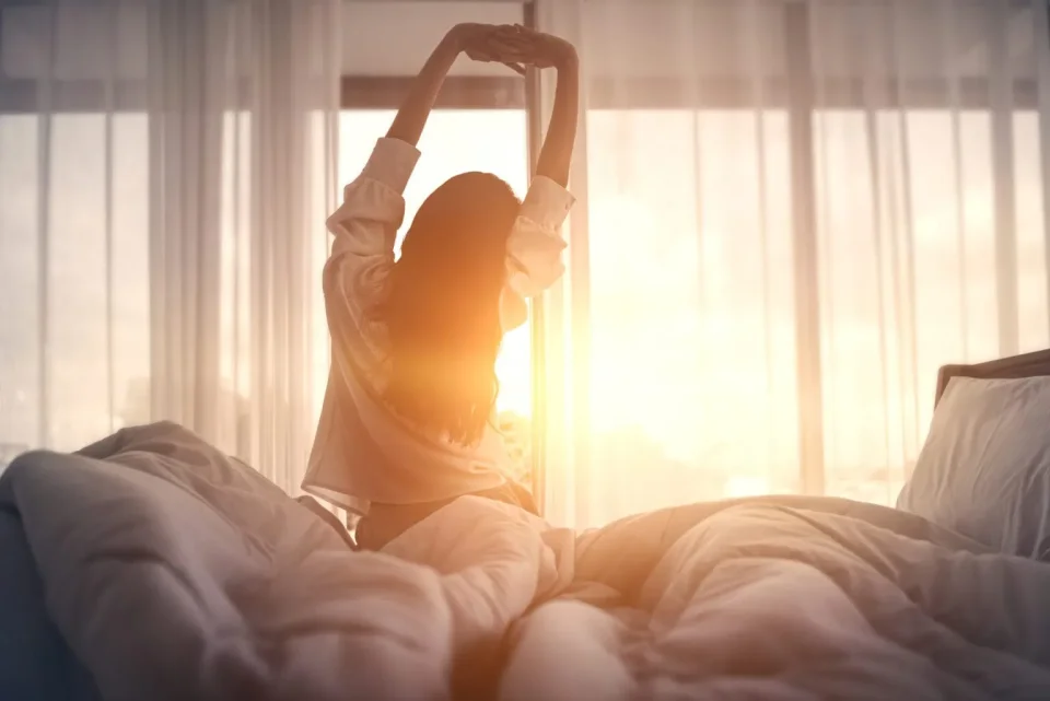 Eine junge Frau sitzt auf der Bettkante und streckt sich in die Morgensonne. 