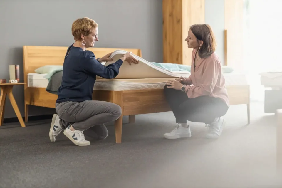 Eine Schlafberaterin der Schlafwerkstatt zeigt einer Kundin den Aufbau einer Matratze mit Unterbett. Beide knien dabei vor einem Massivholzbett und sprechen miteinander.