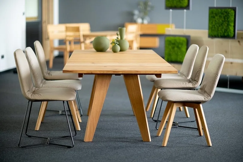 In einer Ausstellung steht ein Esstisch aus hellem Massivholz. Darum sind 6 Stühle mit beigen Lederbezug gestellt. Die Stühle haben abwechselnd Holz- und Metallbeine.