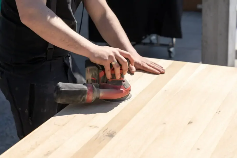Ein Handwerker schleift mit einer Schleifmaschine eine helle Massivholz-Platte.