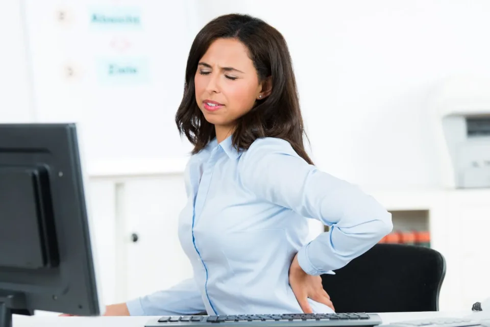Eine Frau mittleren Alters mit längeren dunklen Haaren und einer hellblauen Business-Bluse sitzt im Büro vor einem Monitor. Sie hält sich den Rücken, ihr Gesicht ist vor Schmerzen leicht verzerrt.
