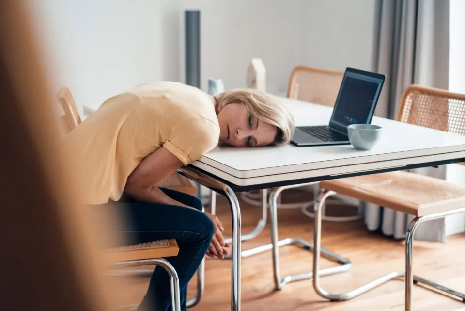 Eine junge Frau ist vor Ihrem Laptop eingeschlafen. Ihr Kopf liegt auf der Tischplatte, der Laptop und eine Teetasse darüber.