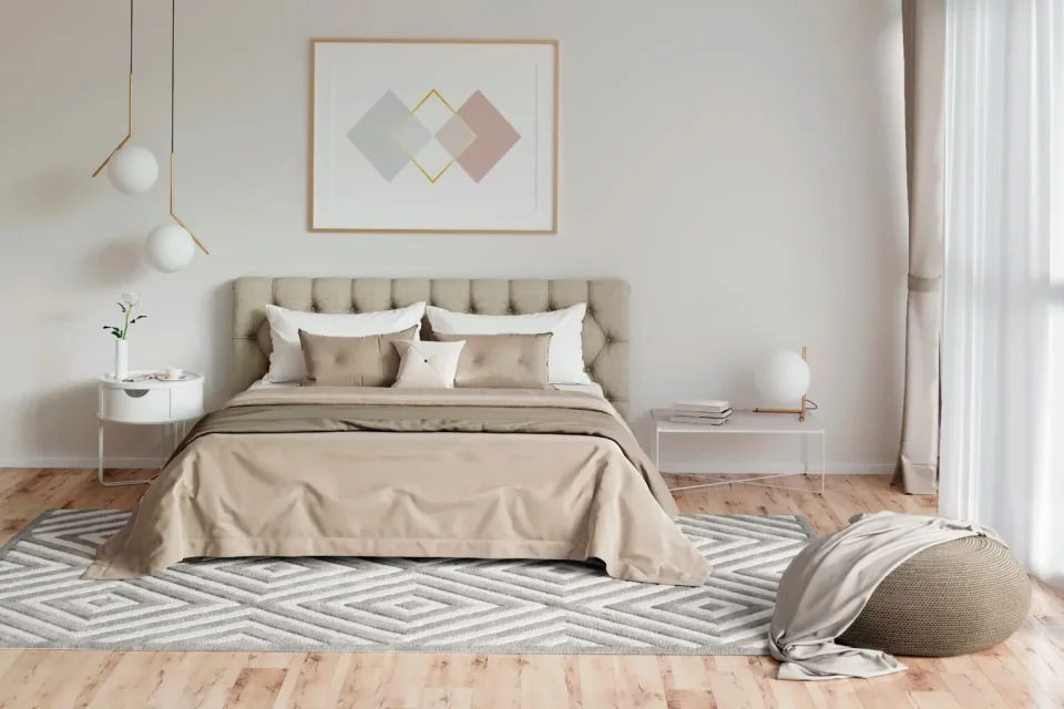 Ein modernes Schlafzimmer in beigen Farbtönen. In der Mitte ist ein großes Doppelbett zu sehen mit einer gepolsterten Rücklehne. Es steht auf einem Parkett und einem in Rauten gemusterten Teppich.