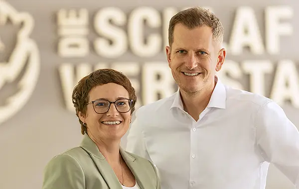 Kathrin und Johannes Rumpfinger stehen nebeneinander und lächeln freundlich in die Kamera. Hinter ihnen an der Wand ist das neue Logo der Schlafwerkstatt mit dem Siebenschläfer zu erkennen.