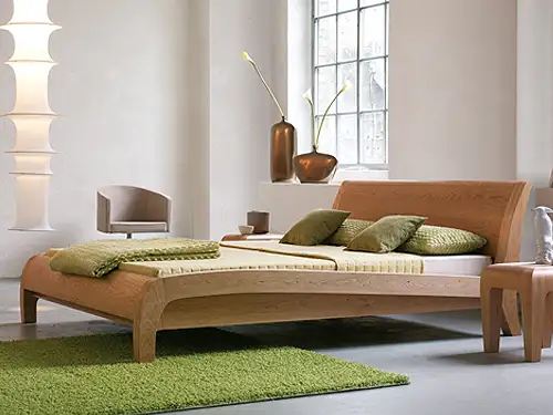 In einem hellen Raum mit einem großen Fenster steht mittig ein Bett. Das Bett hat einen Holzrahmen aus Kirschholz. Vom Fußbereich bis zum Kopfbereich ist der Rahmen leicht geschwungen. Das Bett ist mit einer grünen Decke und grünen Kissen dekoriert. 
