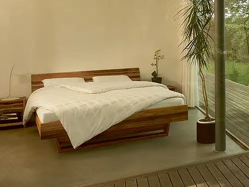 Blick von der Terrasse durch raumhohe Fenster in ein Schlafzimmer mit einem Doppelbett aus Massivholz. Das Bett ist bedeckt mit einer weißen Decke und Kissen. neben dem Bett befinden sich auf beiden Seitenpassende Nachtkästchen.