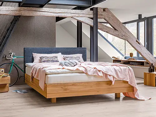 In einem Zimmer mit Dachschräge und Holzbalken befindet sich in der Mitte ein Polsterbett. Das Doppelbett hat einen Rahmen aus hellem Holz und ein gepolstertes Kopfteil mit dunkelblauem Stoff. Auf dem Bett liegt eine rosafarbene Decke und passende Kissen. Neben dem Bett befinden sich zwei passende Nachtkästchen. Sie sind aus demselben hellen Holz. Ein Nachtkästchen hat eine geschlossene Form mit Schubladen, das andere ist wie ein Tisch geformt.