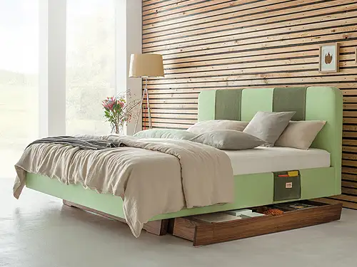 Blick auf ein Doppelbett. Hinter dem Bett befindet sich eine Wand aus hellen Holzlamellen. Rechts sind große Fenster, durch die helles Tageslicht ins Zimmer scheint. Das Bett ist ein Polsterbett. Sowohl der Rahmen als auch das Kopfteil sind weich mit einem hellgrünen Stoff bezogen. Auf dem Bett liegen gemütliche Kissen und eine Decke in passenden Grün und Beigetönen. Unter dem Bett befindet sich eine Schublade aus dunklem Holz, die leicht herausgezogen ist. 