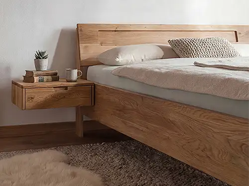 Detailaufnahme eines Betts aus mittelbraunem Massivholz. Die Rückenstütze ist durch eine leichte Einkerbung unterbrochen, das Nachtkästchen direkt am Rahmen befestigt, ohne eigene Beine. Das Bett steht auf einem groben, braunen Teppich mit einem fellartigen Bettvorleger.