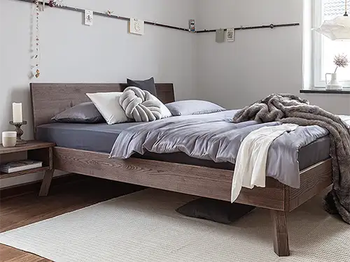 Ein Bett aus dunklem Massivholz in einem schlicht eingerichteten Zimmer. Das Bett hat eine schlichte Form mit einer hölzernen, unverzierten Rückenlehne und vier leicht schräg stehenden Beinen. Das Nachtkästchen ist direkt am Rahmen befestigt, ohne eigene Beine. Auf dem Bett liegen mehrere Decken in verschiedenen Braun- und Grautönen. Auf dem Parkettboden liegt ein dünner weißer Teppich, an der Wand ist eine Fotoleiste zu erkennen.