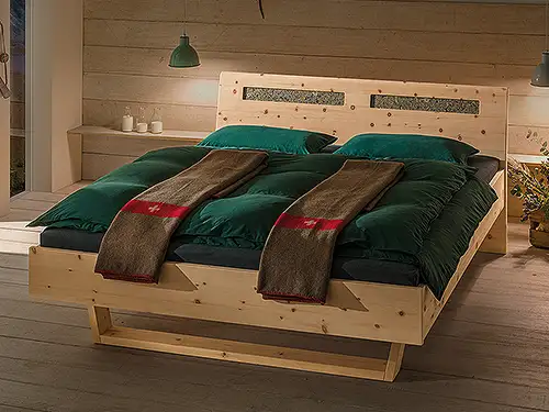 Ein Bett aus hellem Holz in einem modernen alpinen Stil. Die Rückwand ist durch zwei Einkerbungen unterbrochen, in denen Heublumen zu erkennen sind. Das Bett steht in einem abgedunkelten Raum, der an der hinteren Wand mit Holzbrettern verkleidet ist und einen Parkettboden aus dem gleichen Holz hat. Neben dem Bett hängt auf jeder Seite eine kleine dunkelgrüne Lampe von der Decke, als Leselicht. Das Bett ist mit dunkelgrüner Bettwäsche bedeckt. Darauf liegen zwei dunkelbraune Schweiz-Decken mit einem roten Streifen und einem weißen Kreuz.