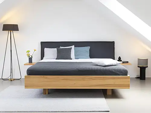 In einem hellen Raum mit Dachschräge steht in der Mitte ein Doppelbett. Das Bett hat einen modernen, geraden Rahmen aus Kernbuche. Der Kopfteil ist mit dunklem Stoff überzogen. Auf dem Bett liegt eine dunkle Decke und Kissen in verschiedenen Blautönen. Die modernen Nachtkästchen sind seitlich am Bett befestigt. Sie sind ebenfalls aus Buchenholz. Neben dem Bett stehen auf beiden Seiten Lampen, die den Raum in warmes Licht tauchen.