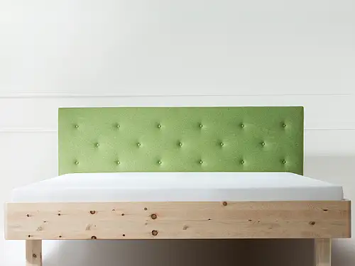 Vor einer weißen Wand mit dezenten Stuckleisten steht ein Doppelbett. Es hat einen geraden Rahmen aus hellem Zirbenholz. Das Bett ist schlicht mit einem weißen Spannbetttuch bezogen. Das Kopfteil ist gepolstert und mit hellgrünem Filz bezogen. 