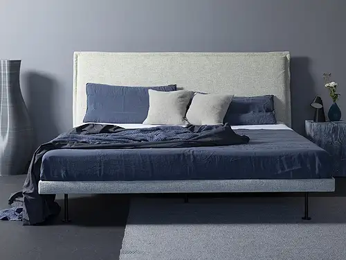 Vor einer graublauen Wand steht ein Doppelbett. Das Bett hat einen dünnen, mit Stoff bezogenen Rahmen in hellem Blaugrau. Das Kopfteil ist ebenfalls bezogen mit einem natürlichen Beigeton. Auf dem Bett liegen eine dunkelblaue Decke und Kissen in verschiedenen Farben. Eine Stehlampe sorgt für eine angenehme Lichtstimmung.
