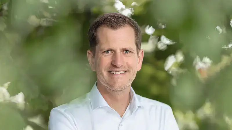 Ein Mann um die 40 mit kurzen braunen Haaren und hoher Stirn. Er trägt einen 3-Tage-Bart und ein weißes Hemd und lächelt freundlich in die Kamera. Hinter ihm sind verschwommen grüne Blätter eines Baums zu erkennen.
