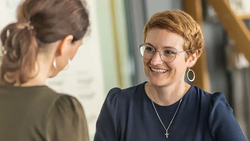 Kathrin Rumpfinger im Gespräch mit einer Kundin, die man nur von hinten erkennt. Sie trägt eine dunkelblaue Bluse und lächelt freundlich.
