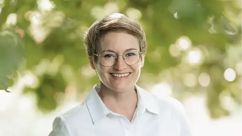 Eine Frau um die 40 mit kurzen rötlichen Haaren. Sie trägt eine Brille mit dünnem Drahtrahmen und eine weiße Bluse und lächelt freundlich in die Kamera. Hinter ihr sind verschwommen grüne Blätter eines Baums zu erkennen.