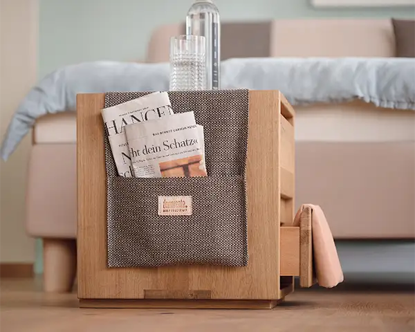 Ein Nachtkästchen aus mittelbraunem Massivholz vor einem beigefarbenen Polsterbett. Seitlich am Nachtkästchen ist eine Stofftasche angebracht, in der eine Zeitung steckt. Auf de Nachtkästchen steht eine Wasserflasche und ein Glas.
