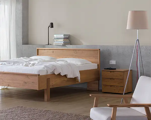 Ein modern und schlicht eingerichtetes Schlafzimmer in skandinavischer Optik. Auf einem Holzparkett steht ein schlichtes Massivholzbett aus mittelbraunem Holz mit weißen Kissen und einer leichten Decke. Daneben ein Nachtkästchen aus dem gleichen Holz, eine moderne Stehlampe auf dünnen Metallbeinen, im Vordergrund ein Polstersessel mit Holz-Armlehne. Auf dem Boden liegt ein graubrauner Teppich. Die sandfarbene Rückwand ist durch einen grauen Absatz geteilt, auf dem ein Leselicht und einige Bücher liegen.