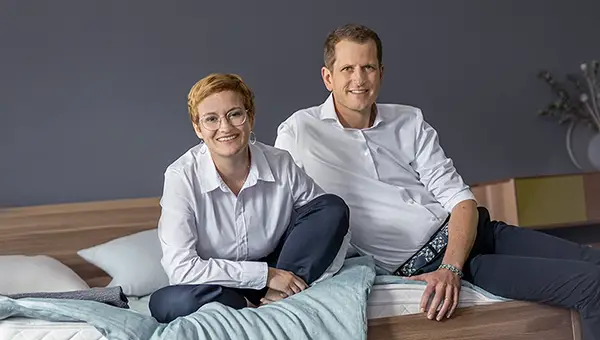 Kathrin und Johannes Rumpfinger. Eine Frau um die 40 mit kurzen rötlichen Haaren, einer dünnen Drahtbrille, und ein Mann um die 40 mit kurzen braunen Haaren und einer hohen Stirn. Beide sitzen nebeneinander auf einem Bett und lächeln freundlich in die Kamera.