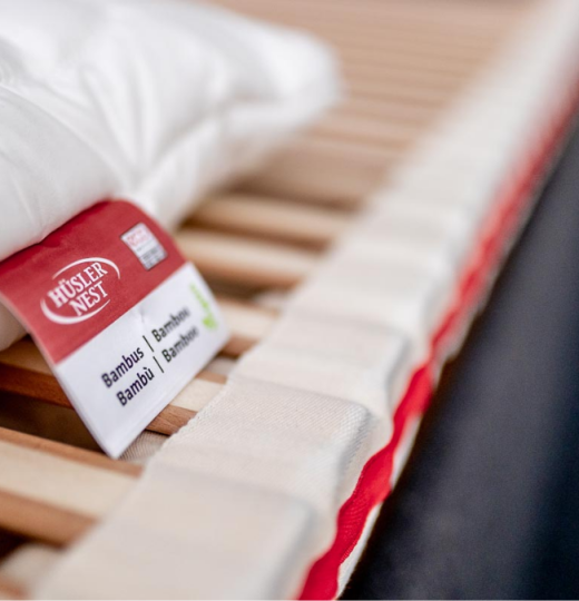 Nahaufnahme eines Hüsler-Nest Federelements. Die Holzlamellen sind weißen Stofftaschen mit einem einem roten Streifen gesteckt. Darauf liegt ein Kissen mit einem Label: Hüsler Nest Bambus